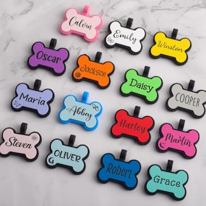 May include: A collection of colorful, bone-shaped pet tags with names like "Calvin," "Emily," and "Winston" printed on them. Each tag has a black loop for attachment and is set against a marble background.