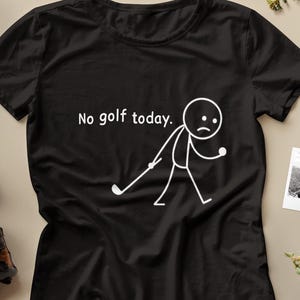 以下が含まれることがあります： 「No golf today.」のテキストと、悲しい顔をしたゴルファーの白い棒人間のデザインが特徴の黒いTシャツ。画像には黒いブーツと装飾的な要素が含まれています。