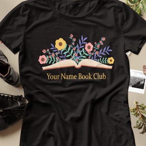 Pode incluir: Uma t-shirt preta de manga curta com um gráfico de um livro aberto de onde saem flores amarelas, cor-de-rosa e roxas e folhas verdes. O texto "Your Name Book Club" está impresso em letras douradas por baixo do gráfico do livro.