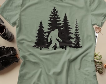 Puede incluir: Una camiseta de manga corta color verde salvia con un estampado gráfico negro de una escena forestal con pinos y la silueta de una criatura Bigfoot caminando por el centro del bosque, centrada en la parte delantera de la prenda.