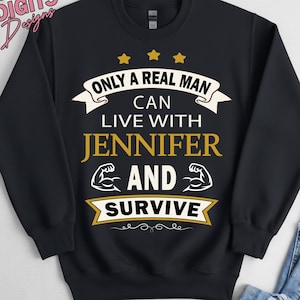 Könnte beinhalten: Ein schwarzes Langarm-Sweatshirt mit Rundhalsausschnitt und weiß-goldener Aufschrift: ONLY A REAL MAN CAN LIVE WITH JENNIFER AND SURVIVE. Das Design enthält drei goldene Sterne über dem Text und zwei weiße Symbole für Bizeps.