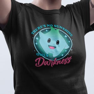 Puede incluir: Camiseta negra con un diseño de dibujos animados. El diseño presenta un personaje azul y verde con una cara sonriente, rodeado por un óvalo y estrellas turquesas. El texto dice "THERE'S NO SUNSHINE ONLY DARKNESS".