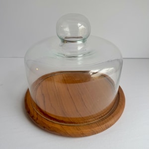 May include: A clear glass cloche with a wooden base. The cloche has a rounded top and a smooth, clear glass body. The base is made of natural wood with a light brown colour.