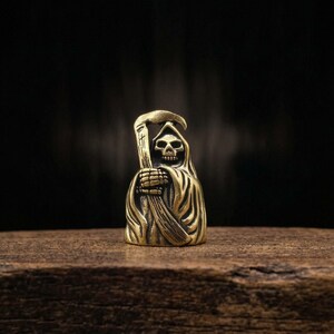 May include: A small, brass-colored figurine of the Grim Reaper, holding a scythe. The figure has a skull face and is draped in a hooded cloak. The figurine is set on a wooden surface against a dark background.