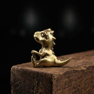 May include: A small, brass-colored dinosaur figurine in a seated yoga pose. The dinosaur has a detailed, textured surface and is positioned on a dark wooden block. The background is a blurred black.
