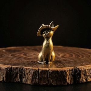 Puede incluir: Pequeña figura de gato en tono latón, con sombrero de vaquero. El gato está sentado con los ojos cerrados y el sombrero ligeramente inclinado. La figura se encuentra sobre una superficie redonda de madera, con fondo oscuro.