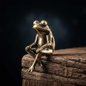 May include: A small, antique-style brass frog figurine with a weathered, dark gold finish. The frog is depicted in a seated, upright position with its front legs resting on its knees and its hind legs dangling downward, showcasing detailed texture.