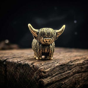 Könnte beinhalten: Kleine, detaillierte Messingfigur einer Highland-Kuh. Die Kuh hat langes, zotteliges Fell, gebogene Hörner und eine kleine Nase. Sie steht auf einer verwitterten Holzoberfläche vor einem dunklen Hintergrund. Die Figur ist etwa 5 cm hoch.