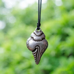 Könnte beinhalten: Ein silberfarbener Muschelanhänger, der an einer schwarzen Kordel hängt. Der Anhänger hat eine detaillierte, strukturierte Oberfläche und ein stilisiertes Design. Der Hintergrund ist ein weiches, verschwommenes Grün.