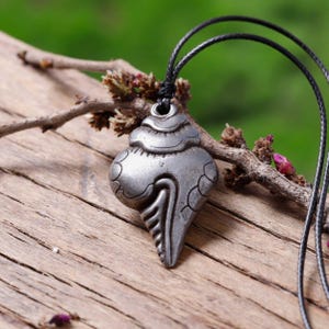 Peut inclure: Un collier avec un pendentif en forme de coquillage argenté sur un cordon noir. Le pendentif est orné de sculptures complexes et d'une pointe pointue.