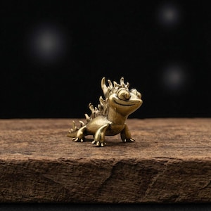 May include: A small, brass-colored lizard figurine with a smiling expression. The figurine has detailed features, including a spiky back and a playful pose. It is set on a wooden surface against a dark background.