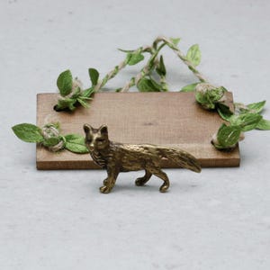 Puede incluir: Una figura de zorro de color bronce se encuentra frente a un pequeño letrero rectangular de madera. El letrero está decorado con hojas verdes y cuerda. El zorro es detallado y parece ser de metal.