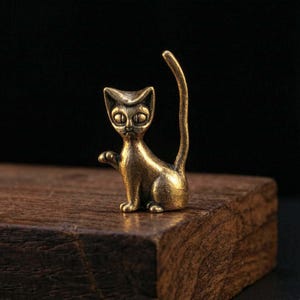 May include: A small, bronze cat figurine with a raised paw and a curved tail. The cat has large, oval eyes and is sitting on a wooden surface. The background is a dark, solid colour.