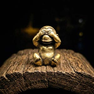 May include: A small, gold-colored monkey figurine in a seated position, covering its eyes with its hands. The figurine is on a piece of weathered wood, with a dark background. The monkey's facial expression is neutral.