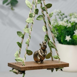 May include: A miniature decorative swing with a small brass-colored animal figurine. The swing is made of a wooden plank and is suspended by twine wrapped with green leaves. The background includes a white pot with white flowers.