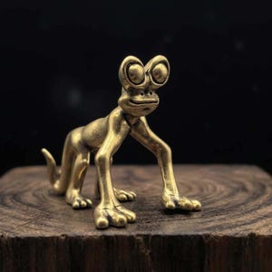 Può includere: Una statuetta in ottone di una lucertola stilizzata con grandi occhi rotondi e arti allungati. La scultura ha una finitura metallica liscia ed è posizionata su un blocco di legno su uno sfondo scuro. I lineamenti della lucertola sono dettagliati, conferendole un aspetto stravagante.