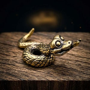 May include: A detailed brass snake figurine with a textured body and a curled pose. The snake has a detailed head with open eyes and a protruding tongue. The figurine is set against a dark background and a wooden surface.