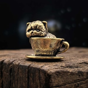 Peut inclure: Petite sculpture en laiton représentant un chien assis dans une tasse à thé. Le chien a une expression renfrognée. La tasse et la soucoupe sont également en laiton, posées sur une surface en bois. Fond sombre.