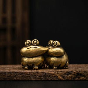 Könnte beinhalten: Zwei kleine, goldene Froschfiguren stehen eng beieinander auf einer Holzoberfläche. Die Frösche haben große, runde Augen und berühren sich mit den Nasen. Der dunkle Hintergrund betont den metallischen Glanz der Frösche.