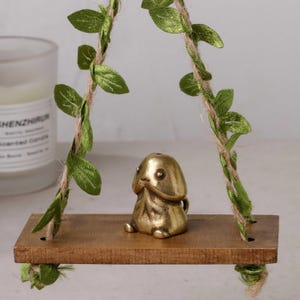 Puede incluir: Una pequeña figura de color latón de una figura estilizada se sienta en un columpio de madera. El columpio está suspendido por una cuerda y adornado con hojas verdes. Una vela perfumada es visible en el fondo.