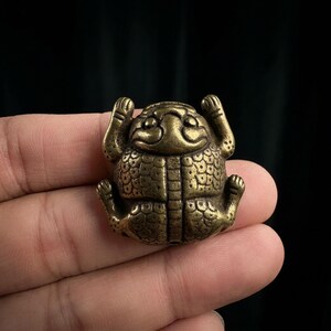May include: A small, brass-colored metal figurine shaped like a stylized frog or toad. The object features a rounded body with a textured, scale-like pattern across its surface, raised front limbs, and bent back legs, all finished in a metallic gold tone.