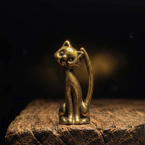 May include: A small, brass cat figurine with a seated pose and an arched tail. The cat has a detailed face and is set against a dark background. The figurine is made of a gold-colored metal and sits on a wooden surface.