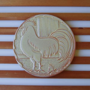 May include: A circular ceramic tile featuring a raised relief design of a rooster standing in profile. The tile has a light yellow glaze with reddish-brown accents highlighting the embossed details of the bird and the background fence structure.
