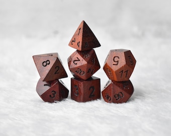 May include: A set of seven wooden polyhedral dice, in various shapes including a d4, d6, d8, d10, d12, d20, and a cube. The dice are a dark reddish-brown color with white numbering. The dice are arranged on a white, textured surface.