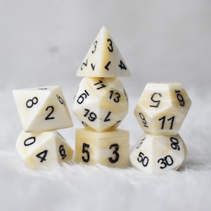 May include: An assortment of ivory-coloured polyhedral dice with black numbering, arranged in a stacked composition. The set includes various shapes: a pyramid, a cube, and several multi-sided dice. The dice are set against a soft, white background.