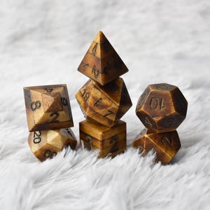 Peut inclure: Un ensemble de six dés bruns avec des chiffres blancs, dont un dé à quatre faces, un dé à six faces, un dé à huit faces, un dé à dix faces, un dé à douze faces et un dé à vingt faces. Les dés sont posés sur une surface blanche et duveteuse.