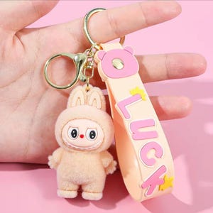 May include: A beige bunny keychain with a gold-colored clasp and a strap that says "LUCK" in pink letters. The bunny has a cute, cartoonish design with a smiling face and a fluffy texture. The strap is light orange with a pink bear head.