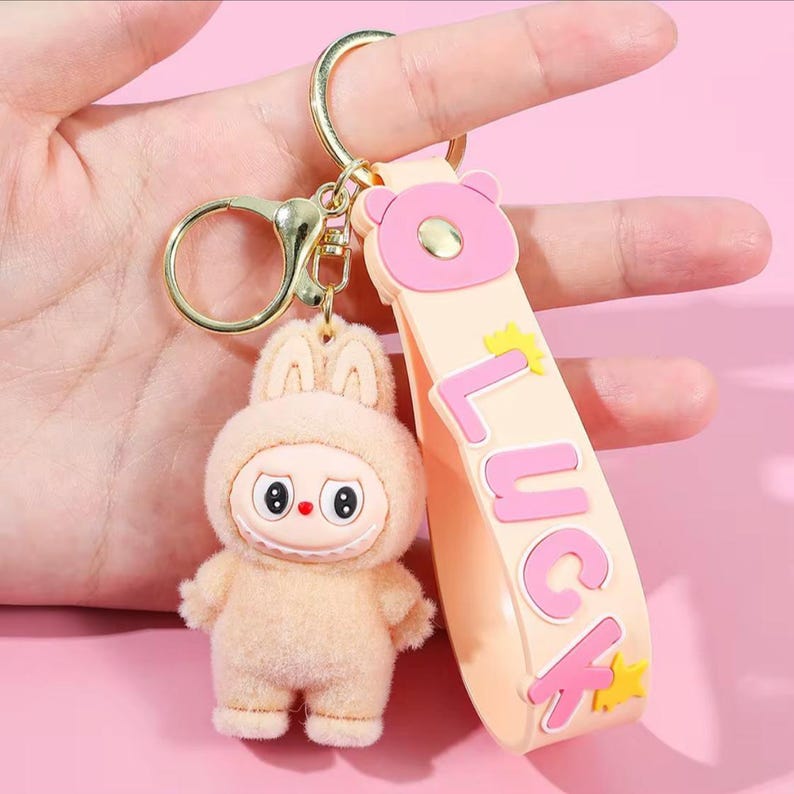 May include: A beige bunny keychain with a gold-colored clasp and a strap that says "LUCK" in pink letters. The bunny has a cute, cartoonish design with a smiling face and a fluffy texture. The strap is light orange with a pink bear head.