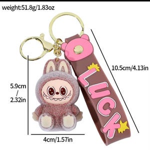 May include: A novelty keychain featuring a cartoon character in a bunny suit. The keychain has a brown strap with the word "LUCK" in pink. The character is 2.32 inches tall and 1.57 inches wide. The strap is 4.13 inches long.