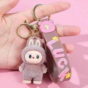 May include: A keychain featuring a plush, cartoon-like figure in a bunny suit with a stern expression. The keychain includes a brown strap with the word "LUCK" in pink letters and gold stars, and a gold-colored clasp.