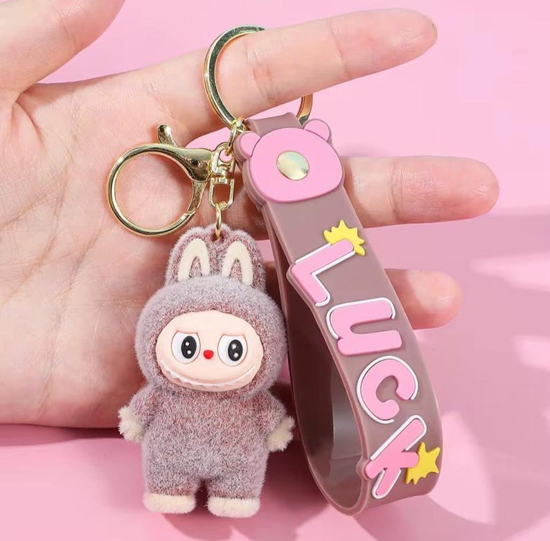 May include: A keychain featuring a plush, cartoon-like figure in a bunny suit with a stern expression. The keychain includes a brown strap with the word "LUCK" in pink letters and gold stars, and a gold-colored clasp.