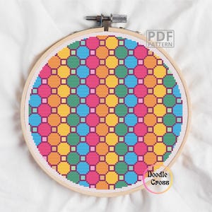 Può includere: Un colorato motivo a punto croce in un telaio da ricamo in legno. Il design presenta un motivo ripetuto di cerchi e quadrati nei toni del rosa, arancione, giallo, verde e blu. I testi "PDF Pattern" e "Doodle Cross" sono visibili.