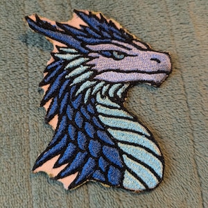 May include: An embroidered patch featuring the profile of a dragon head. The design uses shades of dark blue, light blue, and lavender thread with black outlines. The dragon has sharp, pointed crests along the back of its head and neck.