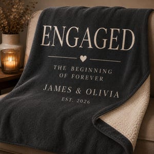 May include: A dark charcoal gray throw blanket with a cream-colored sherpa lining. The front features the text ENGAGED, a small heart icon, THE BEGINNING OF FOREVER, the names JAMES & OLIVIA, and the text EST. 2026 printed in a light cream color.