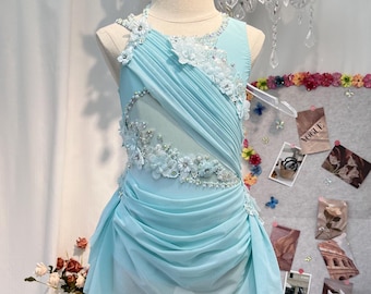May include: A light blue sleeveless dress on a mannequin, featuring diagonal pleated fabric, a sheer mesh panel, and intricate floral appliques with silver beads and sequins along the neckline and side, creating a layered, flowing skirt silhouette.