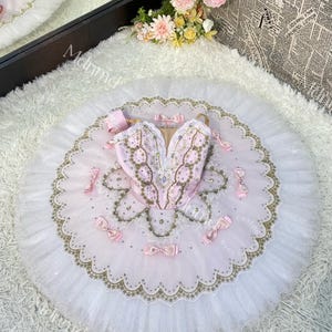 Może przedstawiać: Różowa i biała balerina z ozdobnymi złotymi wykończeniami. Balerina ma różowy gorset z kwiatowym wzorem i białą spódnicę z tiulu.