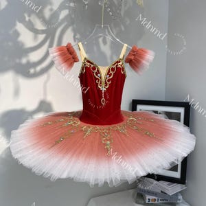 Könnte beinhalten: Ein rotes Samt-Ballett-Tutu mit goldener Stickerei und einem mehrschichtigen Ombre-Rock, der von Pfirsich zu Weiß übergeht. Das Oberteil hat goldene Verzierungen und Strasssteine, mit passenden Armstulpen.