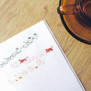 以下が含まれることがあります： ページに様々な色の動物のステッカーが貼られた白いノート。ステッカーには恐竜、猫、ガチョウなどが含まれています。