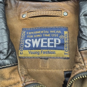 Pu&ograve; includere: Una giacca in pelle marrone con colletto in pelle nera e un'etichetta rettangolare in tessuto blu con il testo SWEEP, EXPERIMENTAL WEAR FOR LONG TIME USE e Young Fashion. La giacca presenta una cerniera in metallo e cuciture visibili.