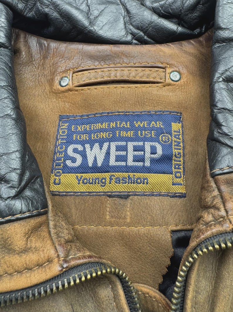 Pu&ograve; includere: Una giacca in pelle marrone con colletto in pelle nera e un'etichetta rettangolare in tessuto blu con il testo SWEEP, EXPERIMENTAL WEAR FOR LONG TIME USE e Young Fashion. La giacca presenta una cerniera in metallo e cuciture visibili.