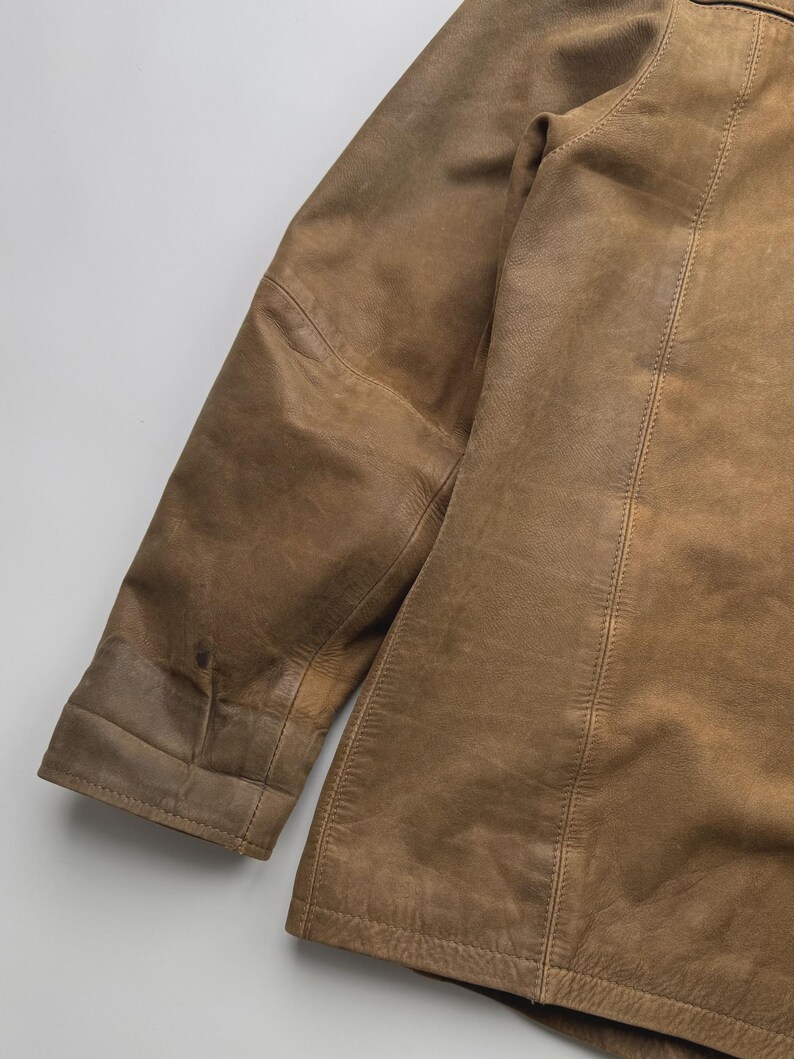 Pu&ograve; includere: Una giacca in pelle color cuoio con maniche lunghe, polsini cuciti e dettagli con cuciture verticali lungo il pannello frontale. Il capo presenta una consistenza liscia e opaca con variazioni naturali sulla superficie in pelle marrone chiaro.