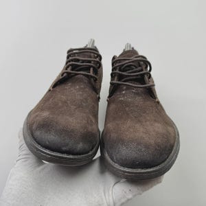 Op de afbeelding: Een paar bruine su&egrave;de enkellaarsjes met donkerbruine veters. De laarzen hebben een ronde neus en een dikke, donkere zool. De schoenen worden gepresenteerd tegen een lichtgrijze achtergrond.