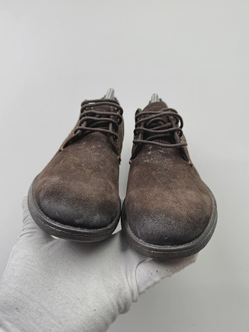 Op de afbeelding: Een paar bruine su&egrave;de enkellaarsjes met donkerbruine veters. De laarzen hebben een ronde neus en een dikke, donkere zool. De schoenen worden gepresenteerd tegen een lichtgrijze achtergrond.