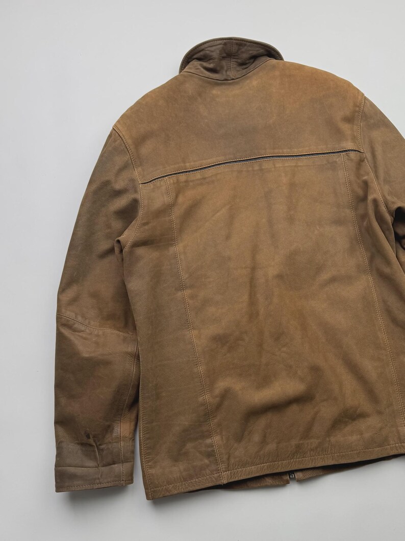 Pu&ograve; includere: Una giacca in camoscio color marrone chiaro con colletto alto, maniche lunghe e una cucitura orizzontale sulla parte superiore della schiena. Il capo presenta dettagli di cuciture visibili lungo i pannelli verticali e una texture liscia e opaca.