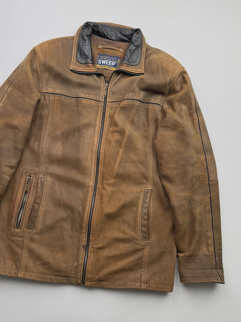 Pu&ograve; includere: Una giacca in pelle marrone con colletto nero e cerniere nere. La giacca presenta una chiusura centrale con zip, due tasche verticali con zip sul davanti e un'etichetta all'interno del colletto con la scritta SWEEP Young Fashion.