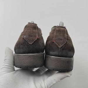 Op de afbeelding: Een paar bruine su&egrave;de schoenen, van achteren bekeken, met een textuurzool. De schoenen hebben een laag profiel en een casual ontwerp. De bovenstukken hebben decoratieve stiksels en kleine accenten van rode en blauwe draad. De schoenen worden vastgehouden door een gehandschoende hand.